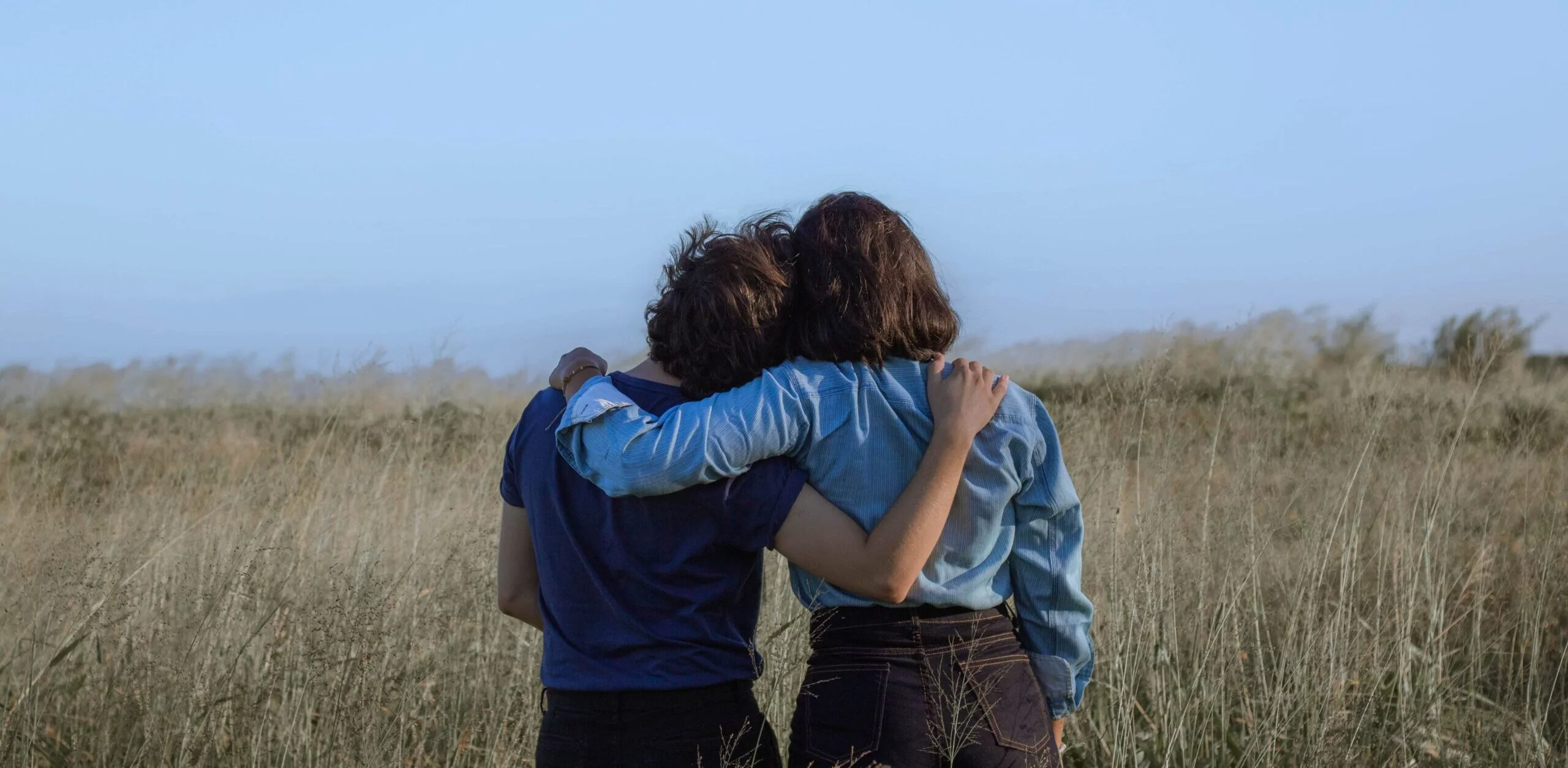 Photo of two friends holding each other by the shoulders from behind, standing in an open field under a blue sky. Cover image for an article titled "Convince your friend to live more sustainably: 9 easy steps"