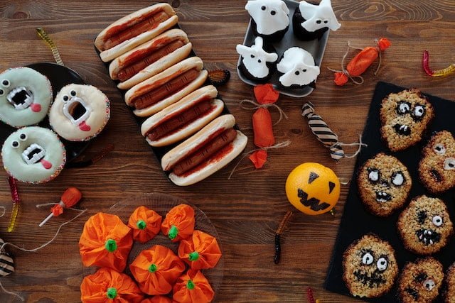 Photo of Halloween-themed treats, including cookies and candies, arranged on a table. Hotdogs are also visible in the photo, possibly indicating a Halloween-themed food spread.