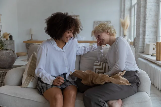 Two women, one White and one Black, sit on a couch with white shirts, smiling and enjoying each other's company.