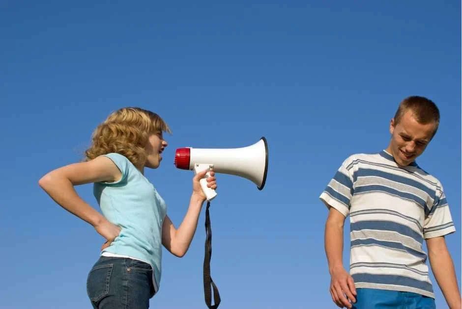 Photo of a girl wearing a t-shirt and jeans holding a hand mic and shouting at a boy also wearing a t-shirt and jeans who appears to be annoyed by the noise pollution.