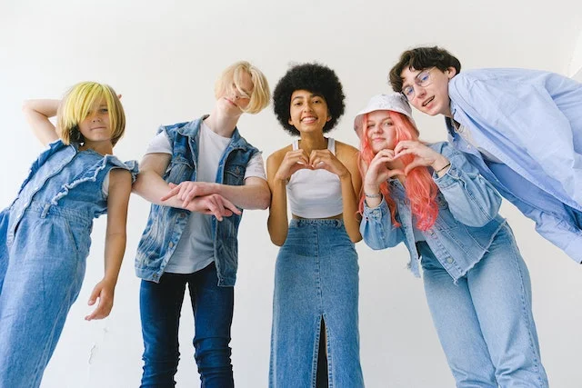 Five multi-racial friends wearing various types of jeans clothing, posing for the camera and making friendly hand gestures.