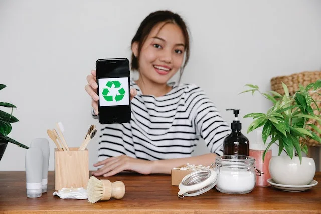 A girl sitting in front of glass pots, containers, potted plants, and a wooden brush stand is holding a smartphone in her right hand and showing it to the viewer. The screen of the phone has a green recycling sign on it, indicating a sustainable lifestyle. The girl is smiling in the photo, suggesting her support for environmental awareness.