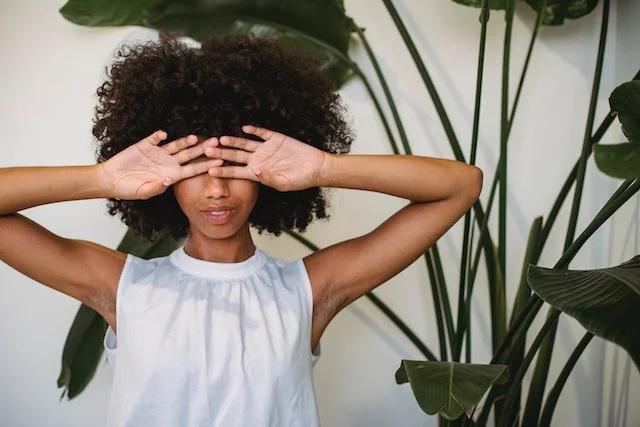 A black woman in a sleeveless white dress stands in front of a white wall with a large house plant partially visible in the background. She covers her eyes with her hands and smiles slightly.