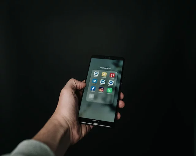 A hand holding a smartphone displaying various social media icons including Facebook, Instagram, Snapchat, WhatsApp, LinkedIn, Pinterest, Twitter, and Telegram.