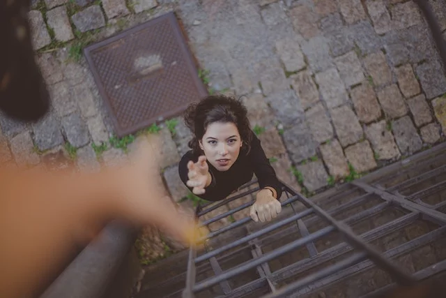 Photo of a hand reaching out from the top of a ladder towards a woman at the bottom of the ladder who is extending her hands to grab the hand that was reached out for her to climb up the ladder.