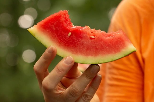 Hand of a person holding a half-eaten watermelon slice. The person is wearing an orange t-shirt, which is slightly visible in the photo."