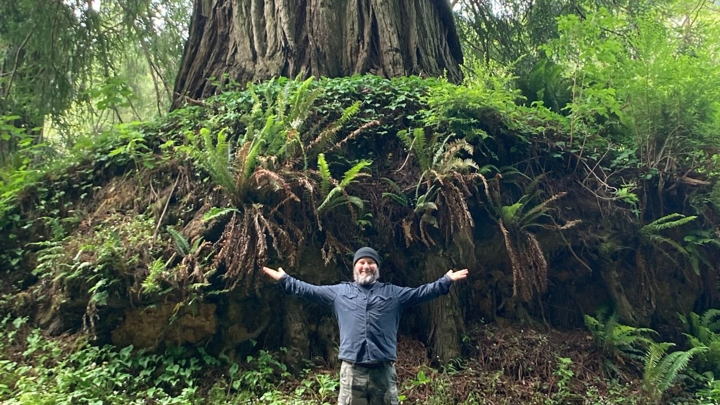 Picture of Rick Frausto standing with his arms wide open in front of a huge tree